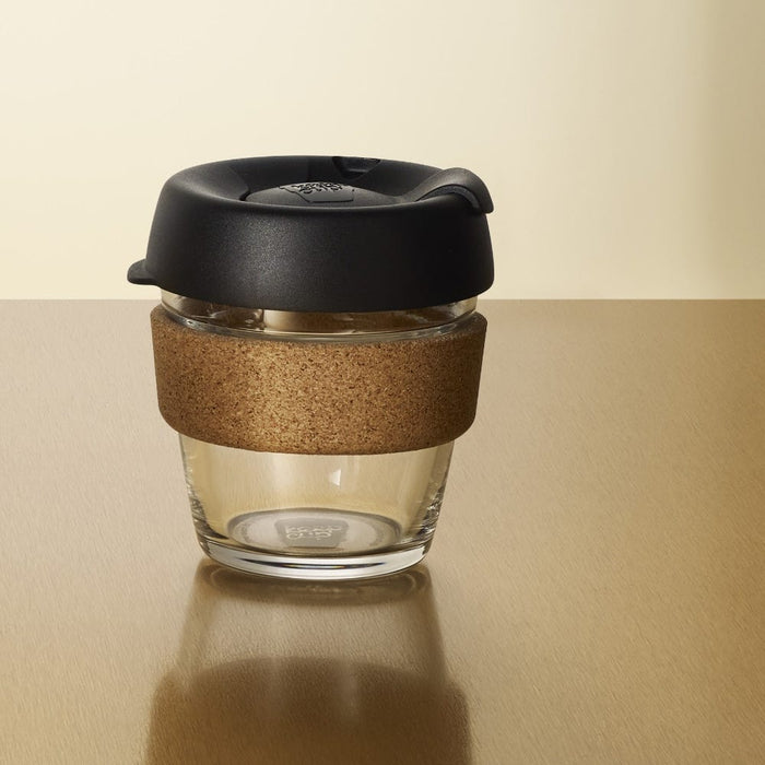 KeepCup Brew Cork XS 6oz in Black, featuring a tempered glass cup with natural cork band and black plastic lid, shown on a wooden surface with a soft beige background.