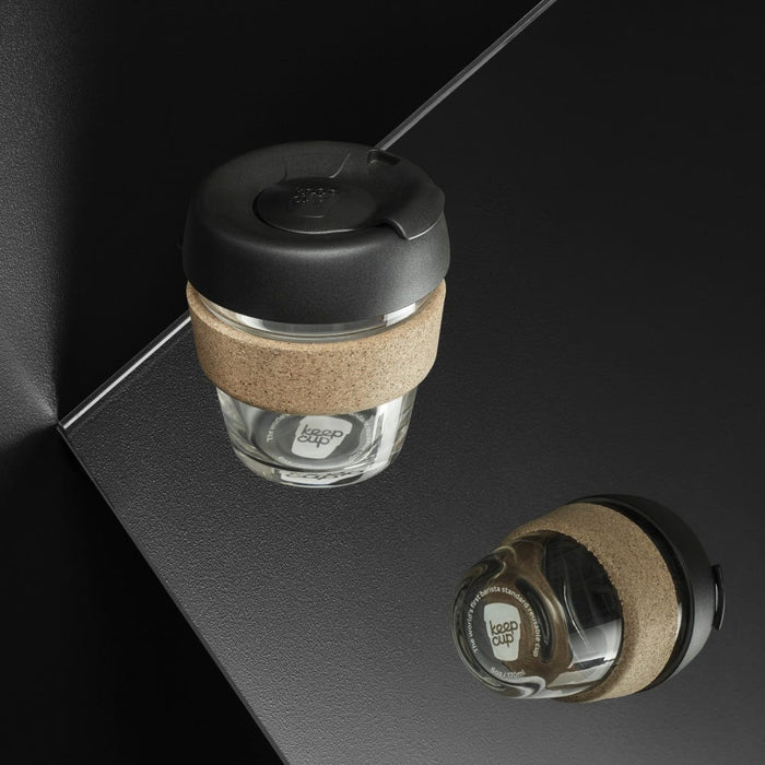 KeepCup Brew Cork XS 6oz in Black, featuring a tempered glass cup with natural cork band and black lid, shown from above on a dark surface with a second cup lying on its side displaying the base and coffee inside.