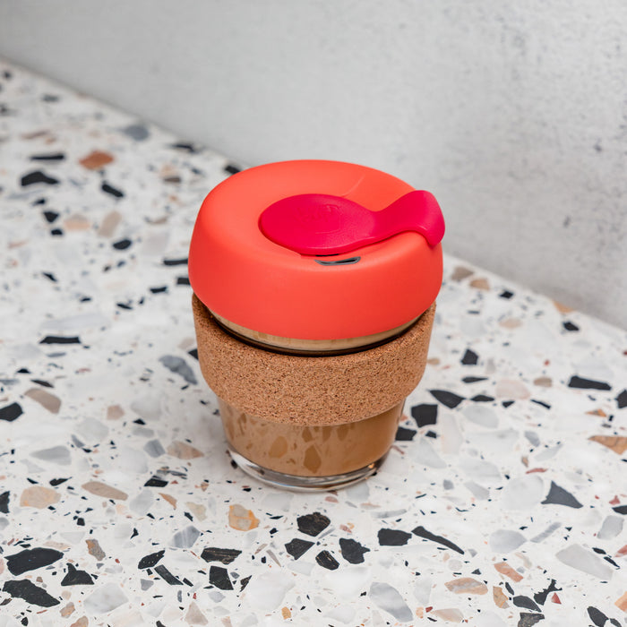 KeepCup Brew Cork XS 6oz in Daybreak colour, featuring a coral-red lid with pink plug, natural cork band, and glass cup containing a latte, placed on a terrazzo surface against a concrete wall.
