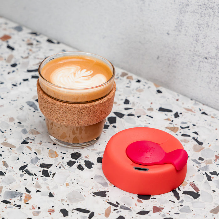 KeepCup Brew Cork XS 6oz in Daybreak colour, filled with a flat white featuring latte art, sits on a terrazzo surface with its coral-red silicone lid placed alongside.