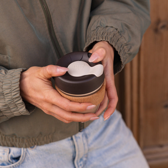 Hands holding a KeepCup Brew Cork S 8oz in Almond, with fingers positioned on the dark brown lid's cream-coloured sipper plug, showcasing the natural cork band against a casual outfit of olive jacket and light denim jeans.