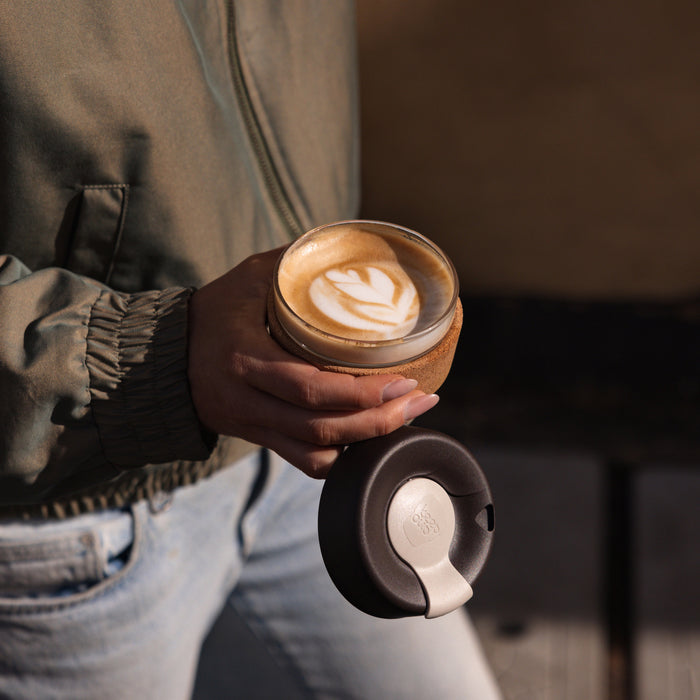 A hand holds an open KeepCup Brew Cork S 8oz in Almond, displaying a latte with leaf-shaped art, while the other hand holds the lid with its cream-coloured closure tab.