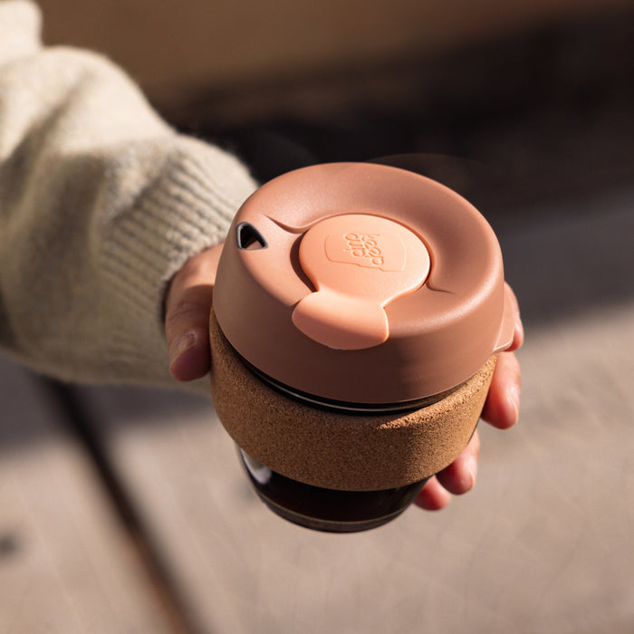 Hand holding a KeepCup Brew Cork S 8oz in Frappe colour, showing the dusty pink lid with plug closure and natural cork band wrapped around the glass cup, photographed from above in warm natural light.