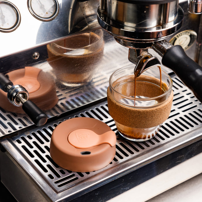 A KeepCup Brew Cork S 8oz in Frappe sits on an espresso machine drip tray, with fresh espresso pouring into the glass cup featuring a cork band, while the terracotta-coloured silicone lid rests beside it.