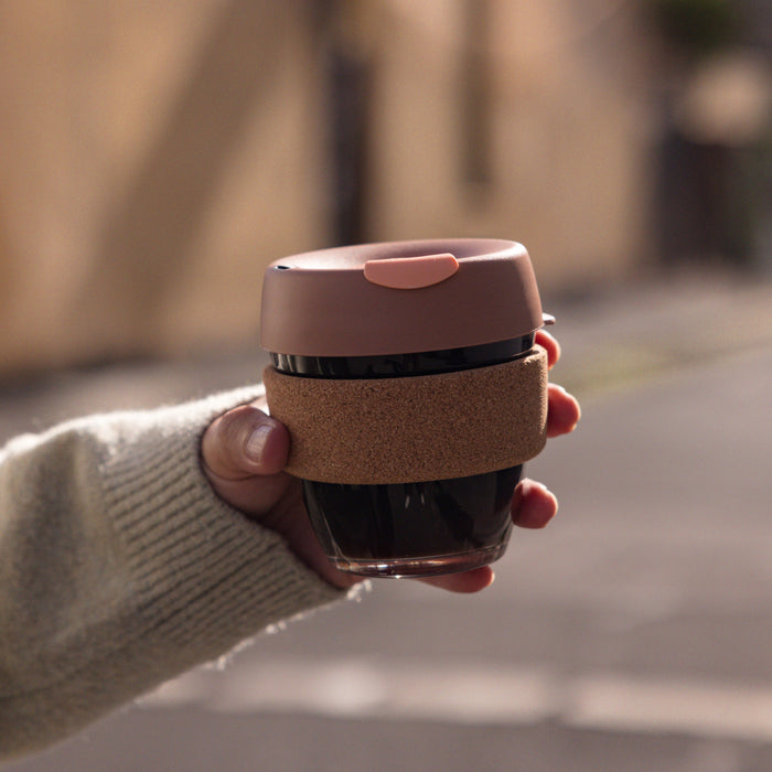 Hand holding a KeepCup Brew Cork S 8oz in Frappe, featuring a dusty pink silicone lid, tempered glass cup filled with dark coffee, and natural cork band, photographed outdoors on a softly blurred street.