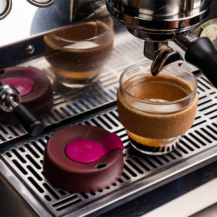 KeepCup Brew Cork S 8oz in Nutmeg colour sits beneath an espresso machine group head, with its burgundy and pink lid resting beside it on the stainless steel drip tray, showcasing the glass cup with natural cork band sleeve.