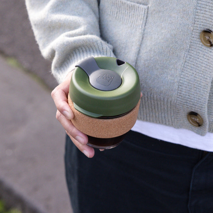 A hand holds the KeepCup Brew Cork S 8oz in Pine, showcasing the olive green lid with grey plug and natural cork band wrapped around the glass cup, with a person wearing a grey cardigan and dark trousers in the background.