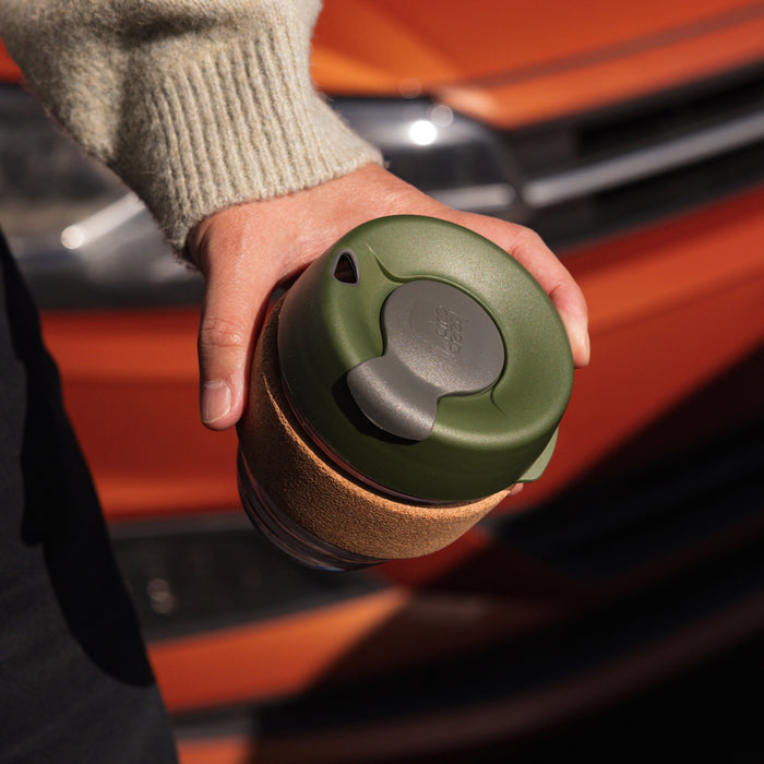A hand holding a KeepCup Brew Cork S 8oz in Pine, viewed from above showing the olive green lid with grey flip-top closure and natural cork band, with an orange vehicle blurred in the background.