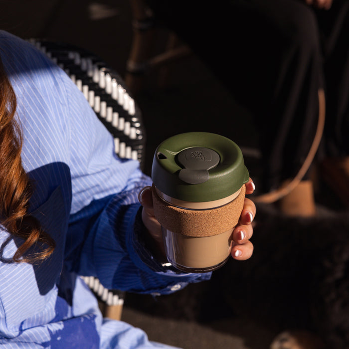 A woman in a blue striped shirt sits in a wicker chair, holding a KeepCup Brew Cork M 12oz in Pine, featuring a dark green lid, cork band and glass cup filled with a creamy coffee drink.
