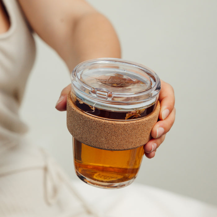 Hand holding a Brew Cork Quicksip M 12oz reusable glass coffee cup filled with tea and loose leaf herbs, featuring a natural cork band sleeve and clear plastic lid, against a neutral background.