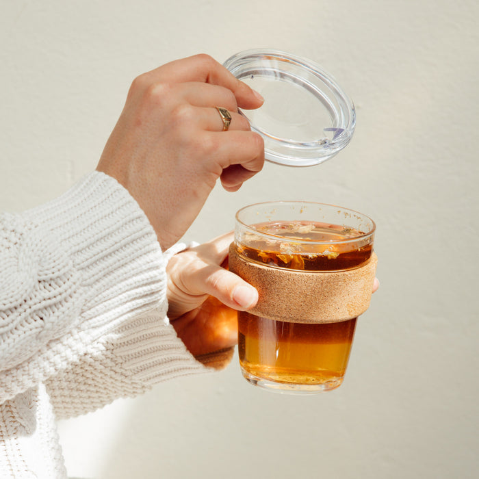 Hands holding a Brew Cork Quicksip M 12oz reusable glass cup filled with golden tea, lifting the clear glass lid to reveal floating tea leaves, with the natural cork band providing grip and insulation.