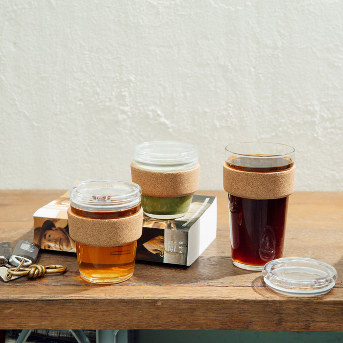Three KeepCup Brew Cork reusable glass cups in different sizes displayed on a wooden table alongside a book and keys, featuring natural cork bands for insulation and clear plastic lids, with the largest cup filled with dark coffee and smaller cups containing lighter beverages.