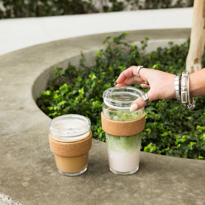 A hand with silver jewellery reaches for the lid of a Brew Cork Quicksip L 16oz KeepCup filled with an iced matcha latte, sitting on a curved concrete ledge beside a smaller cork-banded glass cup containing a creamy coffee drink, with greenery in the background.
