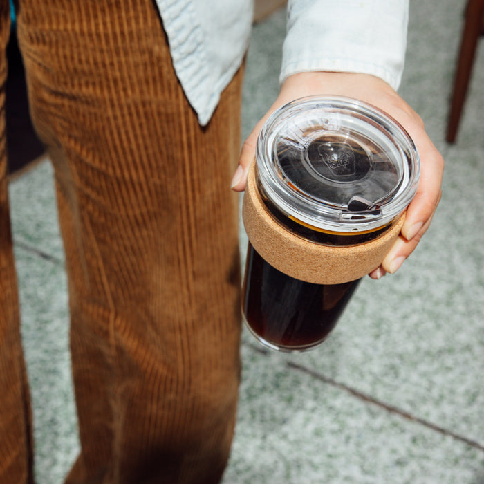 Hand holding a Brew Cork Quicksip L 16oz reusable coffee cup filled with black coffee, featuring a clear glass body with natural cork band and transparent sip lid, photographed from above against a terrazzo floor with brown corduroy trousers visible in the background.
