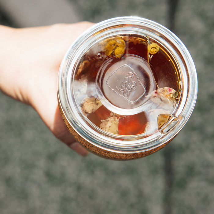 Hand holding a Brew Cork Quicksip L 16oz reusable cup viewed from above, showing the clear plastic lid with flip-top opening and loose leaf tea steeping inside with visible chamomile flowers and rose petals.