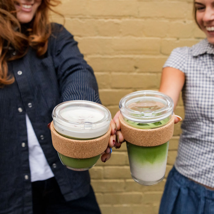 Two smiling women holding KeepCup Brew Cork Quicksip reusable cups filled with layered matcha lattes, featuring cork bands and clear plastic lids, against a yellow brick wall background.