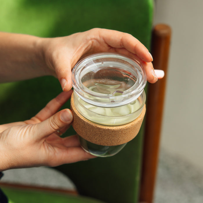 Hands holding a Brew Cork Quicksip S 8oz reusable glass coffee cup with cork band sleeve, filled with pale green tea, viewed from above against a green velvet chair background.