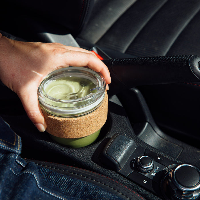 A hand reaches for a KeepCup Brew Cork Quicksip in S 8oz size, filled with a matcha latte, placed in a car cupholder beside the centre console, showcasing the glass cup with cork band in a lifestyle driving context.