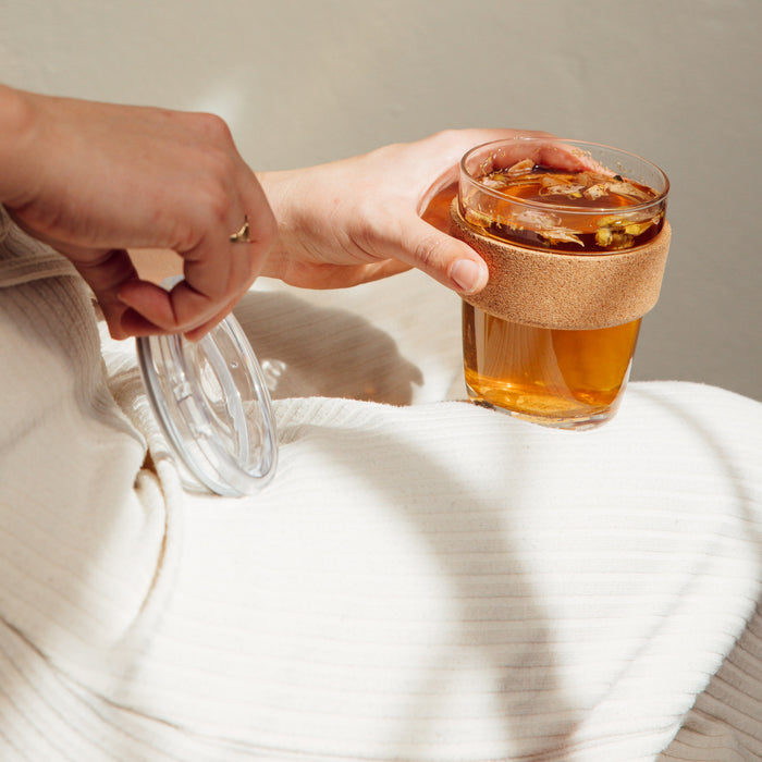 Hand holding a KeepCup Brew Cork Quicksip M 12oz filled with golden tea and floating botanicals, with the clear plastic Quicksip lid held separately, set against a soft cream textured fabric in natural light.