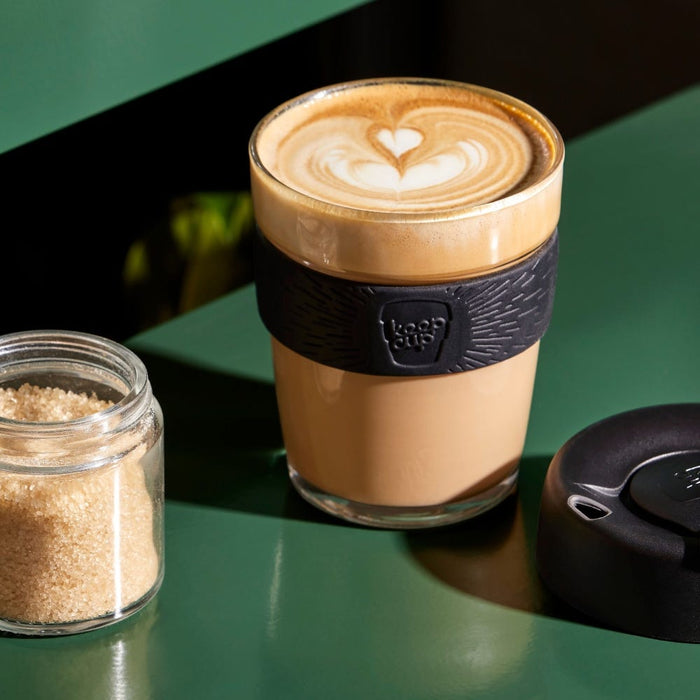 KeepCup Brew M 12oz in Black, filled with a flat white featuring latte art, sits on a green table beside a jar of raw sugar and the black press-fit lid.