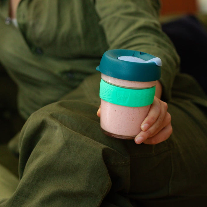 A person in olive green clothing sits holding a KeepCup Brew M 12oz in Eventide, featuring a teal lid, mint green band, and glass cup filled with a pink smoothie.