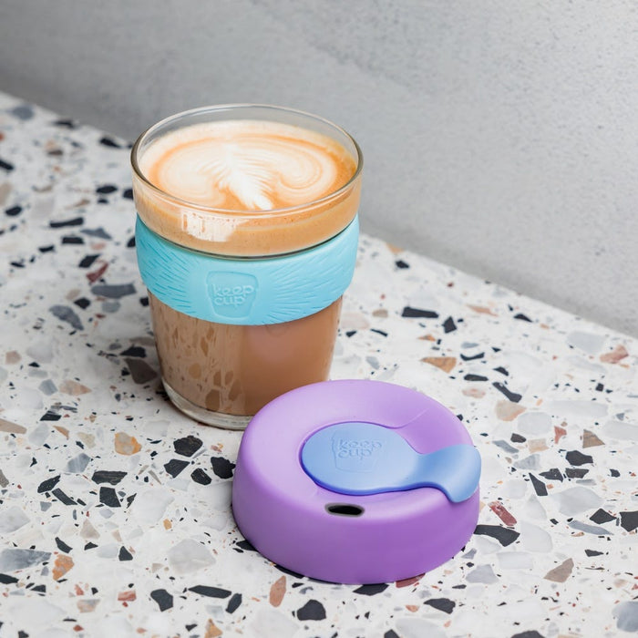 KeepCup Brew M 12oz in Moonlight, a glass reusable coffee cup with turquoise silicone band filled with a latte featuring leaf art, shown with its purple lid placed beside it on a terrazzo surface.