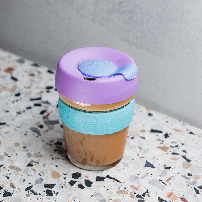 KeepCup Brew M 12oz in Moonlight colour filled with an iced coffee, featuring a lavender lid with blue plug and aqua silicone band, placed on a terrazzo surface against a grey concrete wall.