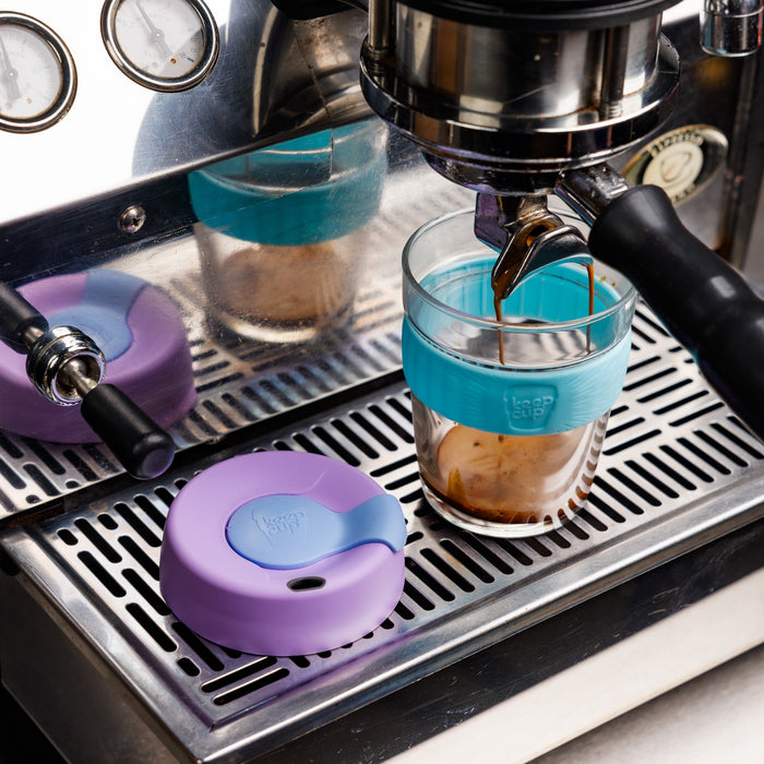 Espresso pouring from a coffee machine into a KeepCup Brew M 12oz in Moonlight, featuring a glass cup with teal silicone band, with the purple and blue lid resting on the stainless steel drip tray beside it.