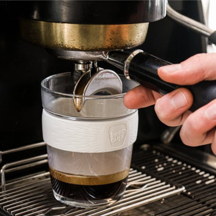 A hand holds a KeepCup Brew M 12oz in Press colourway under an espresso machine portafilter, with fresh espresso pouring into the glass cup featuring a white silicone band.