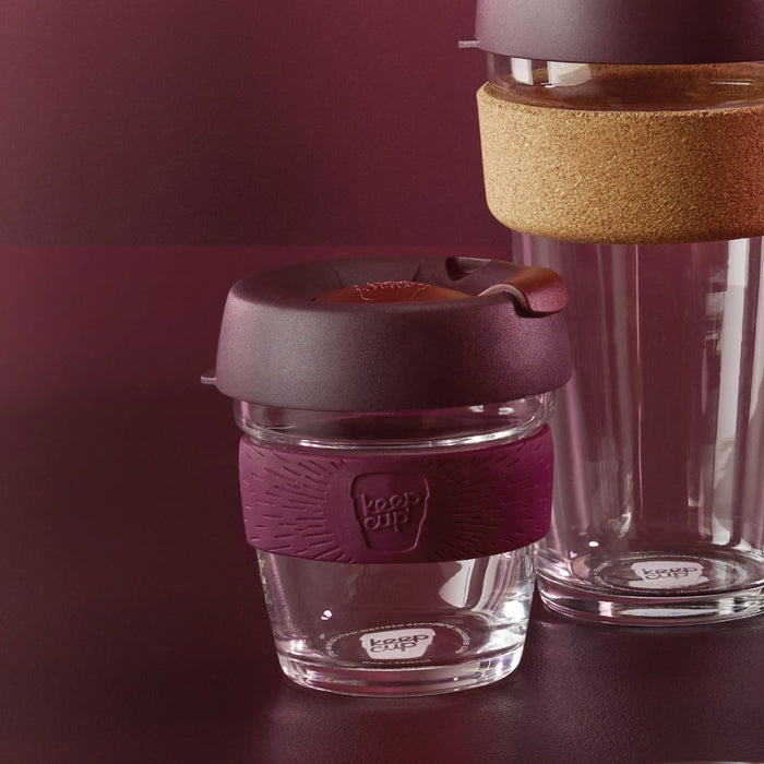 KeepCup Brew XS 6oz in Alder colour, featuring a glass cup with plum-coloured silicone band and dark lid, displayed alongside a larger cork band KeepCup against a rich burgundy background.