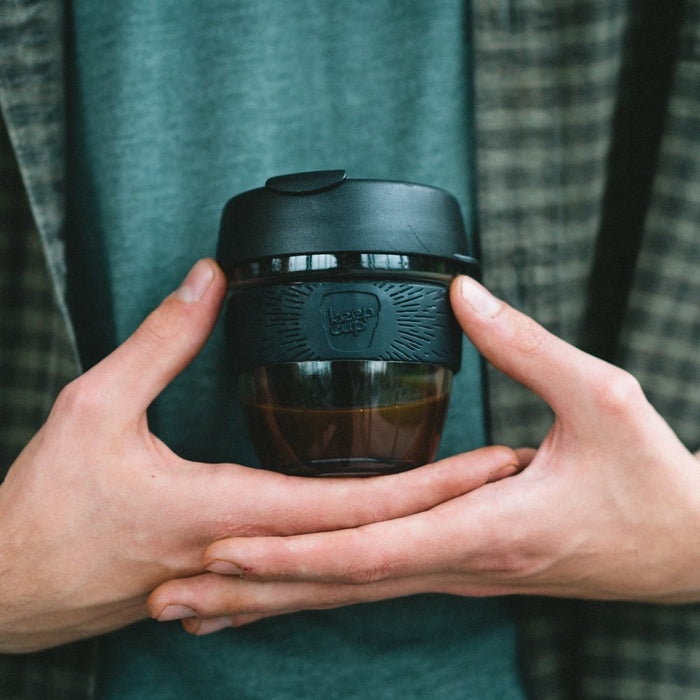 Two hands gently cradling a KeepCup Brew S 8oz in Black, featuring a glass cup with dark liquid inside, black silicone band with embossed KeepCup logo, and black lid, held against a teal fabric background.