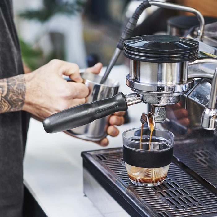 A tattooed barista pulls a double espresso shot into a KeepCup Brew S 8oz in Black, the glass cup with black silicone band sitting on the drip tray of a professional espresso machine.