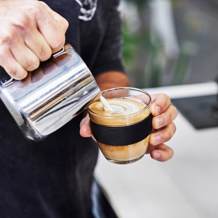 A barista pours steamed milk from a stainless steel pitcher into a KeepCup Brew S 8oz in Black, creating latte art in the glass reusable cup with its signature black silicone band.