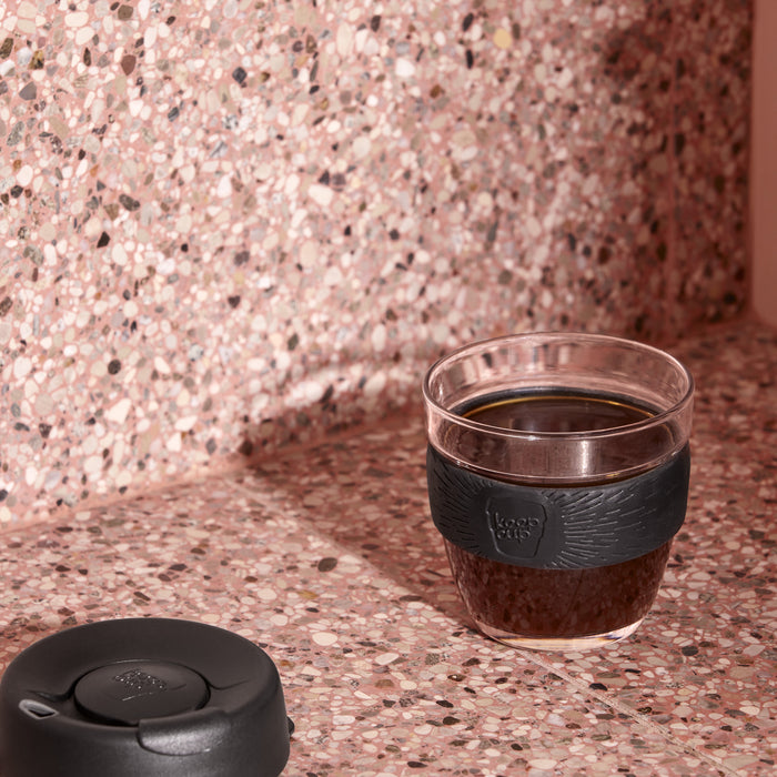 KeepCup Brew S 8oz in Black, filled with black coffee with the lid removed, sits on a pink terrazzo benchtop and matching splashback, with the black lid resting beside it.