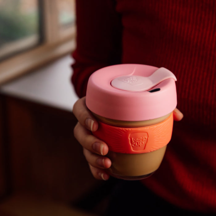 A hand holds a KeepCup Brew S 8oz in Tangerine, featuring a pink silicone lid with flip-top closure, orange silicone band, and tinted glass cup, with a person in a red knit jumper visible in the soft-focus background.