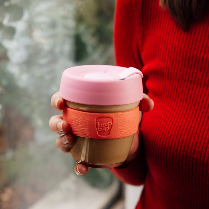 A hand holds a KeepCup Brew S 8oz in Tangerine, featuring a glass cup with coral-coloured silicone band and pink lid, worn by someone in a red knit jumper against a soft, blurred outdoor background.