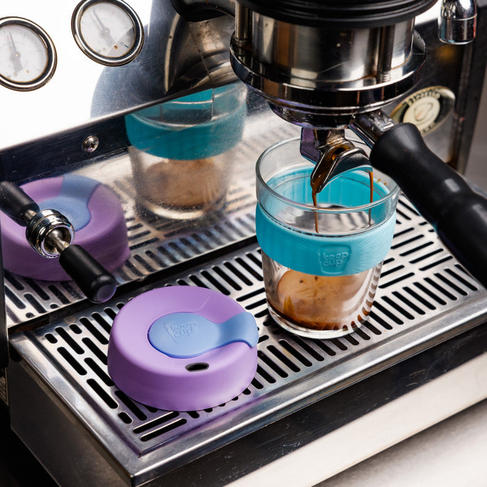 A KeepCup Brew with turquoise silicone band sits on an espresso machine drip tray, with fresh espresso pouring from the portafilter into the glass cup. A purple silicone lid rests beside it on the metal grate.