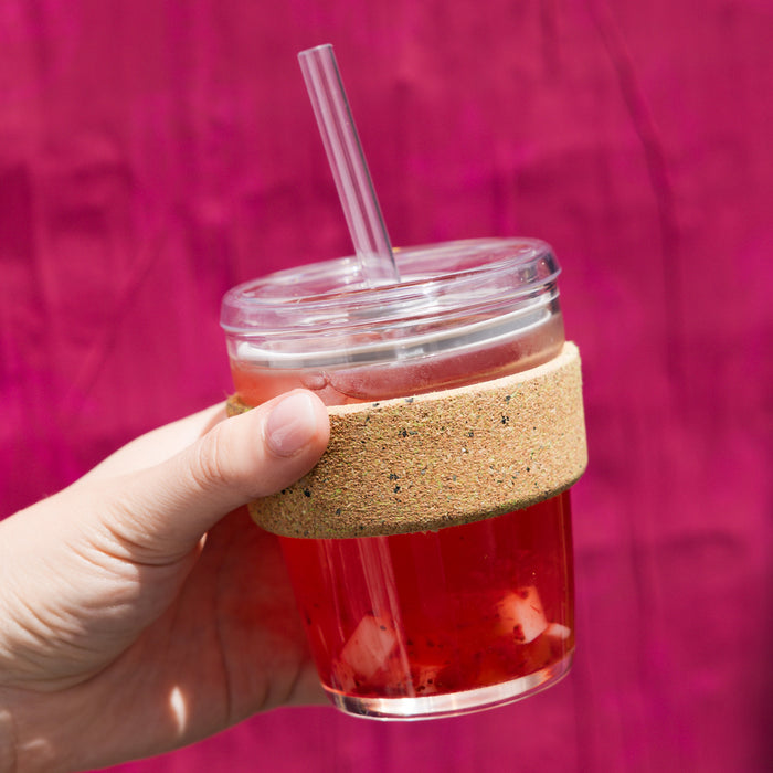 Hand holding a KeepCup Cold Cup Ace M 12oz filled with iced red fruit tea, featuring a clear plastic lid with straw and cork band sleeve, against a vibrant magenta background.