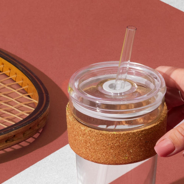 Hand holding a KeepCup Cold Cup Ace M 12oz with cork band and clear plastic lid with plastic straw, positioned on a terracotta tennis court surface next to a vintage wooden tennis racquet.