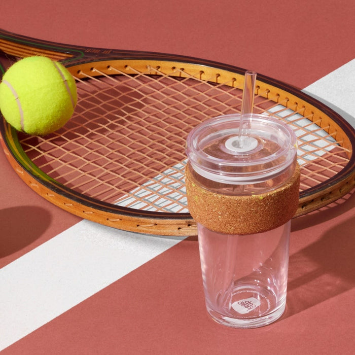 KeepCup Cold Cup Ace L 16oz with cork band and clear straw lid, styled on a terracotta tennis court surface alongside a vintage wooden racquet and yellow tennis ball.