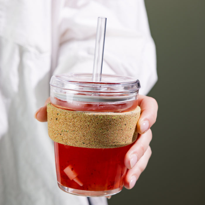 Hand holding a Cold Cup Ace M 12oz filled with iced red fruit tea, featuring a clear lid with plastic straw and natural cork band sleeve, photographed against a muted olive background.