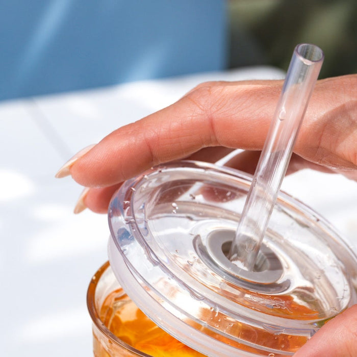 A hand with manicured nails holds a KeepCup Cold Cup Ace filled with iced tea, showcasing the clear reusable straw and transparent lid with condensation droplets in bright outdoor lighting.