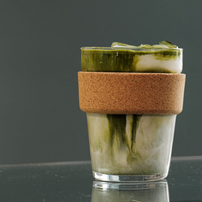 KeepCup Cold Cup Cork M 12oz in Coffee Straw colour, featuring an iced matcha latte with swirled green and white layers, photographed at eye level on a reflective dark surface against a grey background.