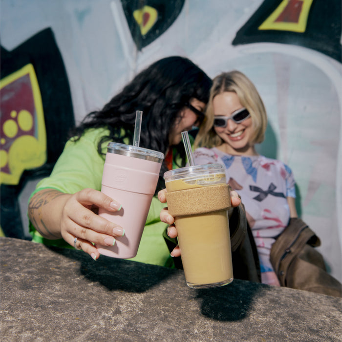 Two friends sitting together at an outdoor table with graffiti in the background, holding up their reusable cups in a cheerful toast—one holding a pink KeepCup and the other holding the Cold Cup Cork L 16oz with Coffee Straw, featuring a glass body with cork band and clear lid with straw.