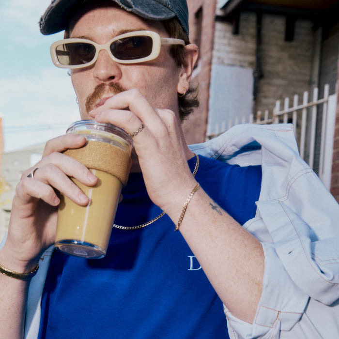 A man wearing sunglasses and a cap sips from a KeepCup Cold Cup Cork L 16oz filled with iced coffee, holding the reusable cup close to his face while carrying a light denim jacket over his shoulder outdoors.