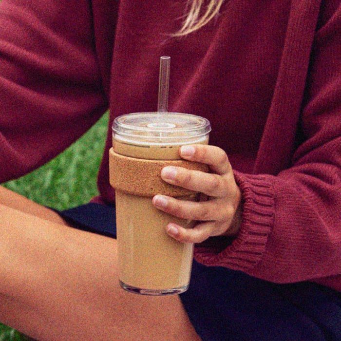 Person in a maroon knit jumper holding a KeepCup Cold Cup Cork L 16oz filled with iced coffee, featuring a cork band sleeve and clear straw, with green plant leaves visible in the background.