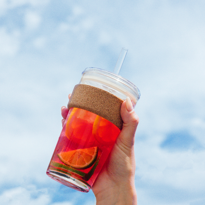 Hand holding a Cold Cup Cork filled with bright red fruit-infused drink and citrus slices against a blue sky with soft clouds, showcasing the cork band grip and clear straw.