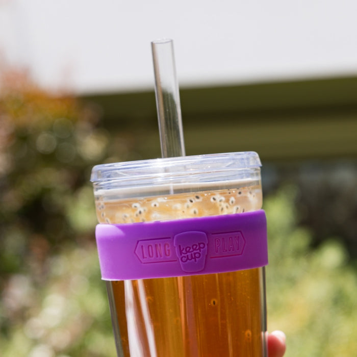 A hand holds the KeepCup Cold Cup Longplay Boba M 12oz in Bubblegum, filled with iced tea and topped with a frothy boba layer, featuring a clear straw and purple silicone band, photographed outdoors with soft garden bokeh in the background.