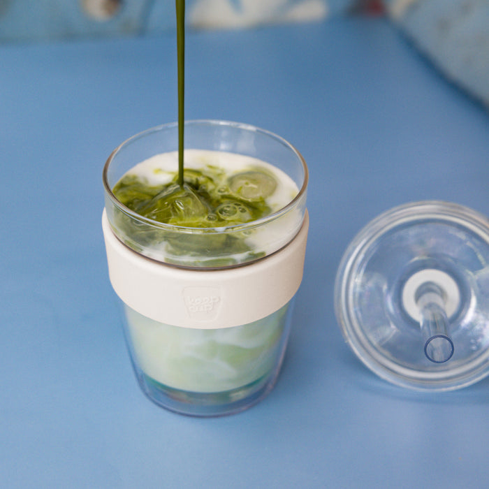 Matcha being poured into a KeepCup Cold Cup Longplay Boba M 12oz in Chalk, showing the layered green tea and milk with ice cubes, the cream-coloured silicone band, and the clear lid with straw hole resting beside it on a blue surface.
