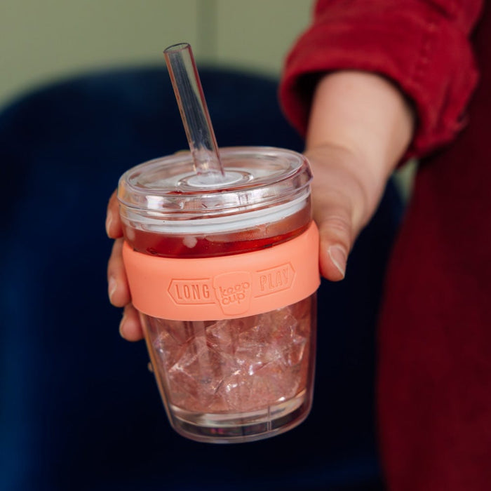 Hand holding a KeepCup Cold Cup Longplay Boba M 12oz in Clay, filled with iced tea and ice cubes, featuring a clear plastic body with coral-coloured silicone band and transparent straw lid.